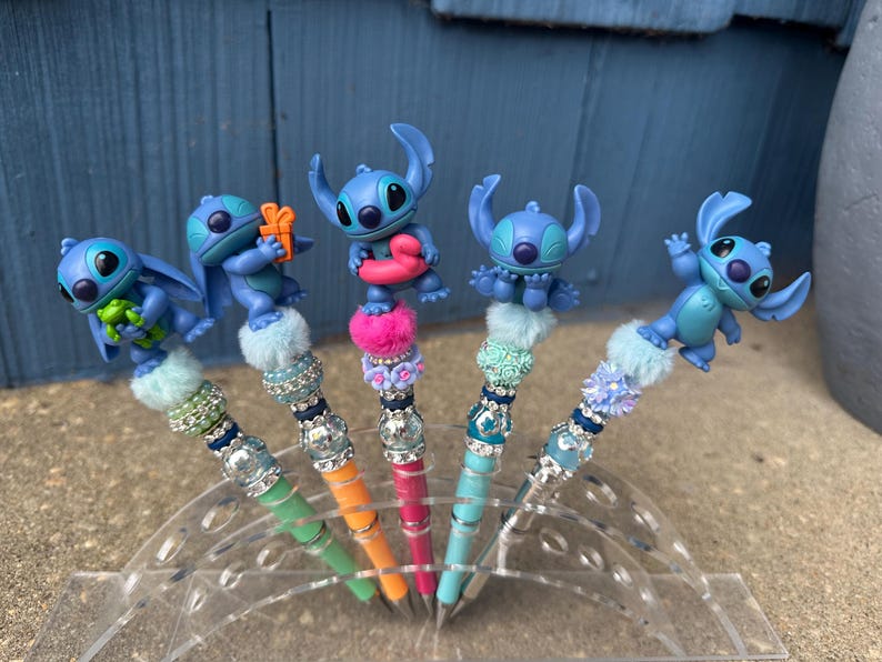 May include: Five decorative pens featuring blue alien character figures. Each pen has a different colored barrel, including green, orange, and pink. The pens are embellished with decorative beads and fluffy accents, and are displayed in a clear plastic holder.