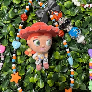 K&ouml;nnte beinhalten: Ein Schl&uuml;sselanh&auml;nger mit Perlen, der eine Jessie-Figur aus Toy Story zeigt. Der Schl&uuml;sselanh&auml;nger enth&auml;lt eine Vielzahl farbenfroher Perlen, darunter orangefarbene Sterne, blaue Herzen und schwarze Schleifen. Der Hintergrund ist ein Bett aus gr&uuml;nen Bl&auml;ttern.
