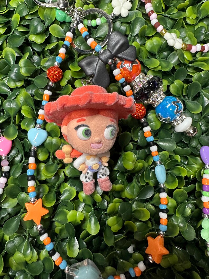 K&ouml;nnte beinhalten: Ein Schl&uuml;sselanh&auml;nger mit Perlen, der eine Jessie-Figur aus Toy Story zeigt. Der Schl&uuml;sselanh&auml;nger enth&auml;lt eine Vielzahl farbenfroher Perlen, darunter orangefarbene Sterne, blaue Herzen und schwarze Schleifen. Der Hintergrund ist ein Bett aus gr&uuml;nen Bl&auml;ttern.