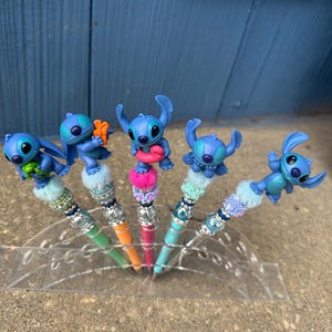 May include: Five pens with blue Stitch character toppers. The pens have colorful barrels in green, orange, pink, and blue. Each pen has a decorative top with beads and faux fur. The pens are displayed in a clear plastic holder.