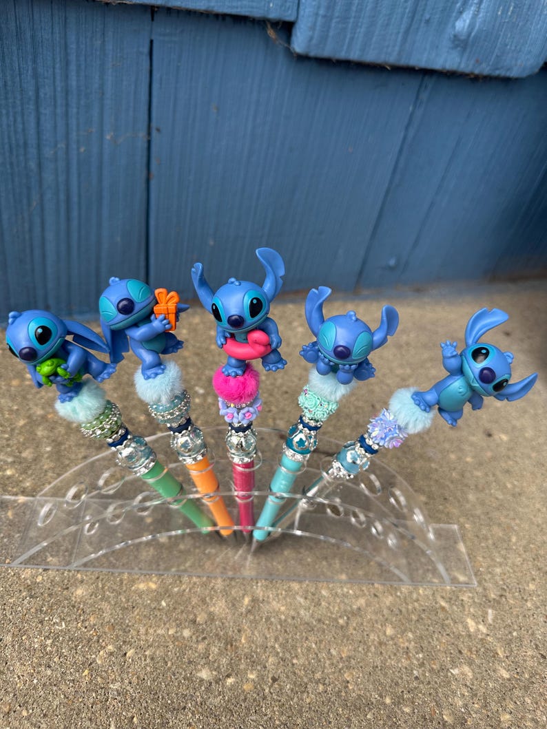 May include: Five pens with blue Stitch character toppers. The pens have colorful barrels in green, orange, pink, and blue. Each pen has a decorative top with beads and faux fur. The pens are displayed in a clear plastic holder.