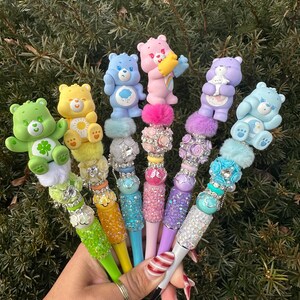 May include: A collection of colorful pens, each topped with a different Care Bear figurine. The pens feature various colors, including green, yellow, blue, pink, and purple, with decorative embellishments like rhinestones and fluffy accents. The pens are held against a backdrop of green foliage.