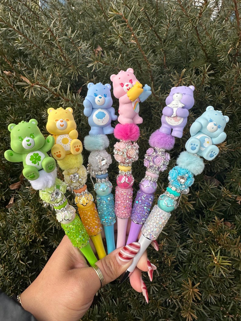 May include: A collection of colorful pens, each topped with a different Care Bear figurine. The pens feature various colors, including green, yellow, blue, pink, and purple, with decorative embellishments like rhinestones and fluffy accents. The pens are held against a backdrop of green foliage.