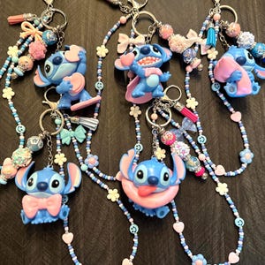Puede incluir: Llaveros con figuras de personajes de dibujos animados azules con detalles rosas. Cada llavero tiene un cord&oacute;n de cuentas en tonos azules, rosas y blancos, adornado con abalorios, lazos y borlas. Los llaveros est&aacute;n sujetos a llaveros plateados.