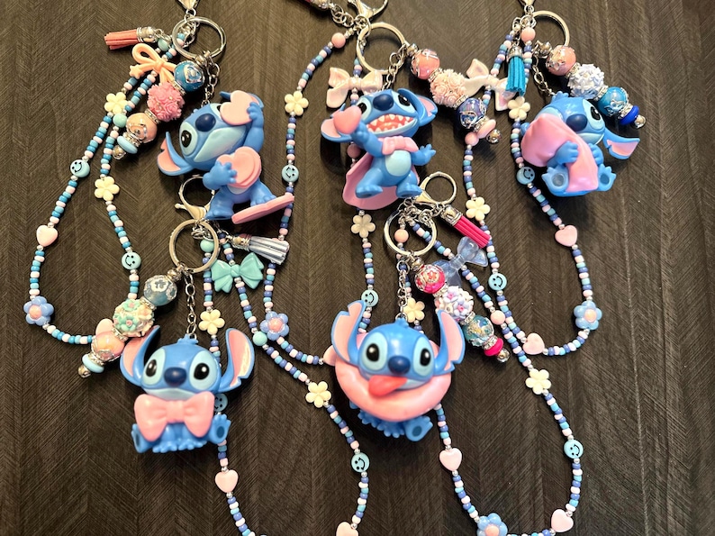 Puede incluir: Llaveros con figuras de personajes de dibujos animados azules con detalles rosas. Cada llavero tiene un cord&oacute;n de cuentas en tonos azules, rosas y blancos, adornado con abalorios, lazos y borlas. Los llaveros est&aacute;n sujetos a llaveros plateados.