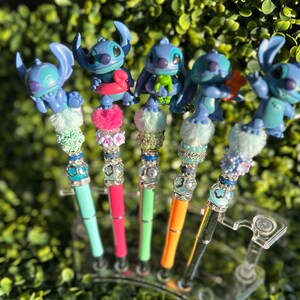 May include: Five colorful pens featuring blue alien figures. Each pen has a different design, with a variety of colors including teal, pink, green, and orange. The pens are decorated with sparkling beads and fluffy accents, creating a whimsical look.