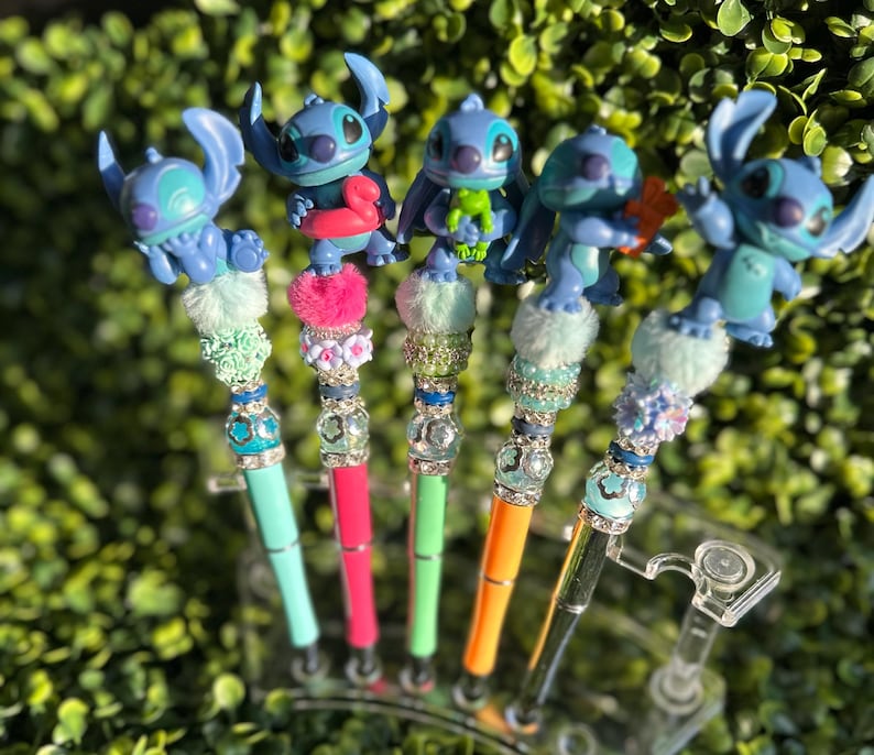 May include: Five colorful pens featuring blue alien figures. Each pen has a different design, with a variety of colors including teal, pink, green, and orange. The pens are decorated with sparkling beads and fluffy accents, creating a whimsical look.