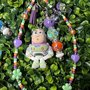 K&ouml;nnte beinhalten: Ein farbenfroher Schl&uuml;sselanh&auml;nger mit einer Buzz Lightyear-Figur. Der Schl&uuml;sselanh&auml;nger enth&auml;lt einen lila Quastenanh&auml;nger, herzf&ouml;rmige Perlen und bl&uuml;tenf&ouml;rmige Perlen. Die Perlen sind in verschiedenen Farben gehalten, darunter Orange, Gr&uuml;n und Lila.