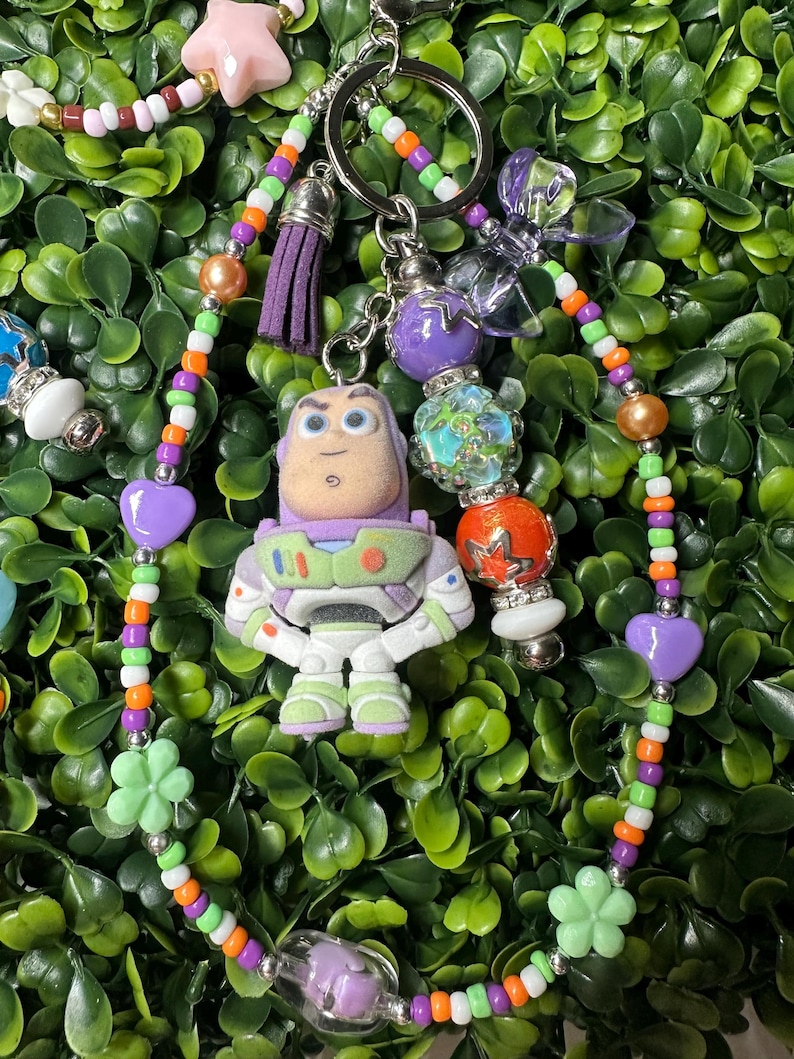 K&ouml;nnte beinhalten: Ein farbenfroher Schl&uuml;sselanh&auml;nger mit einer Buzz Lightyear-Figur. Der Schl&uuml;sselanh&auml;nger enth&auml;lt einen lila Quastenanh&auml;nger, herzf&ouml;rmige Perlen und bl&uuml;tenf&ouml;rmige Perlen. Die Perlen sind in verschiedenen Farben gehalten, darunter Orange, Gr&uuml;n und Lila.