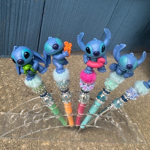 May include: Five decorative pens featuring blue cartoon character figures. Each pen has a different colored barrel, including green, orange, pink, and teal. The pens are displayed in a clear acrylic holder, with a blue wooden background.