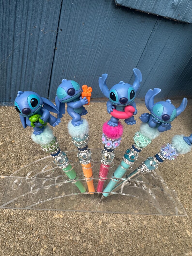 May include: Five decorative pens featuring blue cartoon character figures. Each pen has a different colored barrel, including green, orange, pink, and teal. The pens are displayed in a clear acrylic holder, with a blue wooden background.