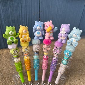 May include: A collection of six colorful pens, each topped with a Care Bear figurine. The pens feature a variety of colors, including green, yellow, blue, pink, purple, and white. The pen barrels are decorated with glitter and beads, and the pens are displayed in a clear plastic stand.