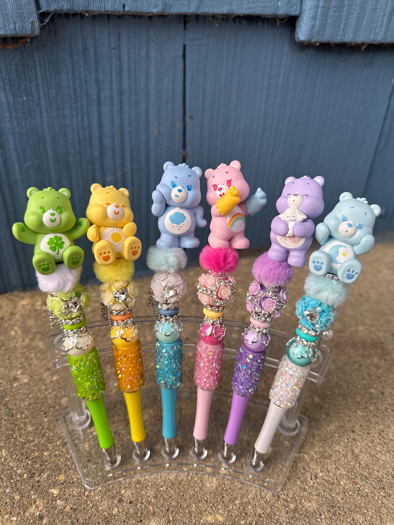 May include: A collection of six colorful pens, each topped with a Care Bear figurine. The pens feature a variety of colors, including green, yellow, blue, pink, purple, and white. The pen barrels are decorated with glitter and beads, and the pens are displayed in a clear plastic stand.