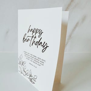 Puede incluir: Una tarjeta de cumplea&ntilde;os blanca con las palabras "happy birthday" en elegante escritura negra. La tarjeta tambi&eacute;n presenta una cita en alem&aacute;n y un delicado dise&ntilde;o floral. La tarjeta est&aacute; doblada y se mantiene en posici&oacute;n vertical.