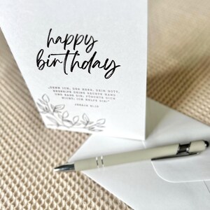 Puede incluir: Una tarjeta de cumplea&ntilde;os blanca con la frase "happy birthday" en escritura negra. Un bol&iacute;grafo y un sobre est&aacute;n junto a la tarjeta. La tarjeta tiene un dise&ntilde;o floral y texto en alem&aacute;n.