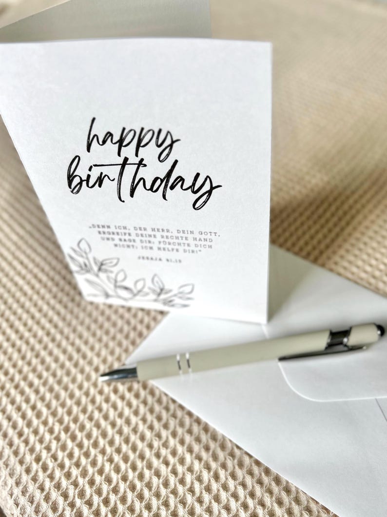 Puede incluir: Una tarjeta de cumplea&ntilde;os blanca con la frase "happy birthday" en escritura negra. Un bol&iacute;grafo y un sobre est&aacute;n junto a la tarjeta. La tarjeta tiene un dise&ntilde;o floral y texto en alem&aacute;n.