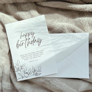 Puede incluir: Una tarjeta de cumplea&ntilde;os blanca con las palabras "happy birthday" en escritura cursiva. La tarjeta tiene un dise&ntilde;o floral y va acompa&ntilde;ada de un sobre blanco. La tarjeta tambi&eacute;n tiene texto en alem&aacute;n.