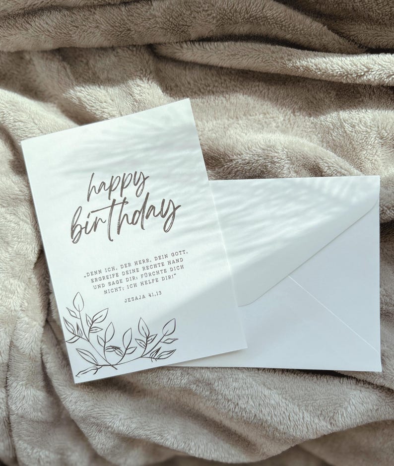 Puede incluir: Una tarjeta de cumplea&ntilde;os blanca con las palabras "happy birthday" en escritura cursiva. La tarjeta tiene un dise&ntilde;o floral y va acompa&ntilde;ada de un sobre blanco. La tarjeta tambi&eacute;n tiene texto en alem&aacute;n.
