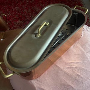 Peut inclure: Une poissonnière ovale en cuivre avec un couvercle en laiton poli et des poignées en laiton assorties de chaque côté. L'intérieur contient un plateau amovible en métal perforé pour la cuisson vapeur, avec une finition métallique robuste.