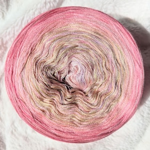 Peut inclure: Une pelote de laine rose, beige et marron avec un effet dégradé. La laine est enroulée en cercle et a une surface texturée.