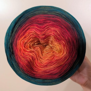 Può includere: Un vivace gomitolo di filato con una sfumatura di colori, che passa dal turchese al rosso e all'arancione al centro. Il filato è ben avvolto, creando una forma circolare. Il filato è tenuto in una mano.