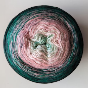 May include: A circular ball of yarn with a gradient colour scheme. The yarn transitions from a dark teal green on the outside to pink and white in the centre. The yarn is tightly wound, creating a smooth, textured surface. Suitable for knitting and crochet projects.