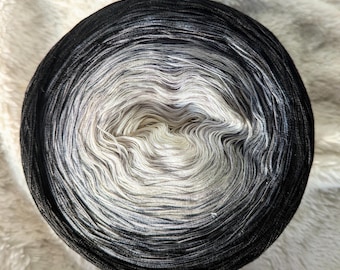 Puede incluir: Ovillo de lana redondo con un degradado de color que va del negro al gris y al blanco. La lana está enrollada firmemente, creando una superficie texturizada. Ideal para proyectos de tejido o ganchillo.