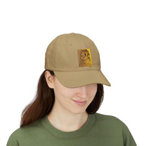 Op de afbeelding: Een beige baseballpet met een rechthoekige patch op het voorpaneel. De patch toont een gedetailleerde, donkerbruine en gouden grafische illustratie van een gestileerd hoofdprofiel met ingewikkelde lijnen en een getextureerde, gouden achtergrond.
