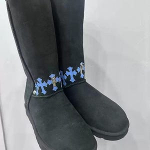 May include: A pair of tall black suede boots featuring a horizontal band of light blue cross-shaped patches around the ankle. Each patch is accented with a small metallic silver cross charm attached to the center, creating a decorative detail.