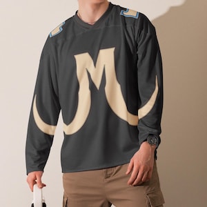 May include: A dark gray long-sleeve jersey featuring a large cream-colored letter M on the chest, light blue accents on the shoulders, and a V-neck collar. The garment is paired with tan cargo pants and a white object held in the hand.