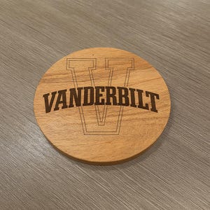 May include: A round wooden coaster with the word "VANDERBILT" in a dark brown font. Above the word is a large letter "V" in a similar font. The coaster is made of light brown wood.