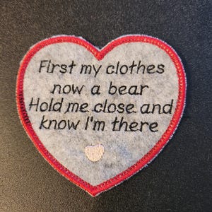 May include: A heart-shaped patch with a grey felt centre and a red stitched border. The patch features embroidered text: "First my clothes now a bear Hold me close and know I'm there." A small pink heart is embroidered at the bottom.