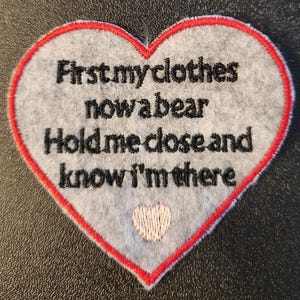 May include: A heart-shaped patch with a grey background and a red border. The patch features the embroidered text "First my clothes now a bear Hold me close and know I'm there." A small, white embroidered heart is at the bottom.
