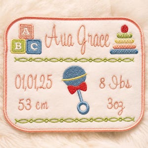 May include: Embroidered baby announcement with the name "Ava Grace", birth date, weight (8 lbs), and length (53 cm). Features a rattle, building blocks, and a stacking toy. The design is on a white background with a pink border.