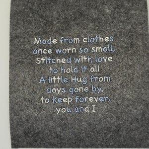 May include: A square piece of dark grey felt fabric featuring light blue embroidered text that reads: Made from clothes once worn so small, Stitched with love to hold it all A little Hug from days gone by, to keep forever, you and I.