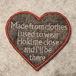 May include: A grey heart-shaped patch with red stitching around the edge. The patch has the embroidered text: "Made from clothes I used to wear Hold me close and I'll be there."