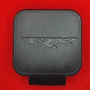 Pode incluir: Uma capa preta retangular com cantos arredondados e as letras "TRX" em relevo em uma fonte estilizada. A capa tem uma pequena aba retangular que se estende da borda inferior. O fundo é vermelho sólido.