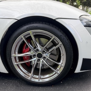 May include: A white sports car front wheel featuring a multi-spoke chrome rim, a black tire with visible SportPlus text, and a bright red brake caliper with white Supra lettering. The car body is white with a black front bumper accent piece.