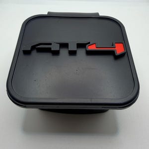 May include: A black rectangular object with the letters "ATT" in a raised design. The "T" and arrow are red. The object has a rounded edge and a small handle on the top.