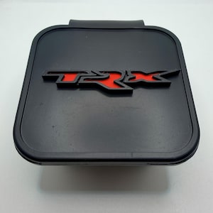 Pode incluir: Peça automotiva retangular preta com as letras vermelhas "TRX" no centro. As letras são contornadas em preto e têm uma aparência em relevo. A peça tem uma borda curva e uma pequena aba na parte superior.