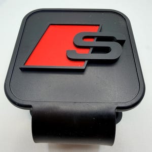 Puede incluir: Emblema de coche negro con fondo rojo y logotipo "S" negro. El emblema es rectangular con esquinas redondeadas y un clip negro curvado en la parte inferior.