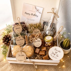 May include: A gift box filled with tea, cookies, honey, and candies. The box includes tea varieties like Jamaica, Fruity Paradise, Chamomile, and Maui Mango. A card reads "Happy Birthday" and a coaster says "Keep calm and Drink Tea".