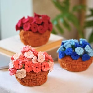 May include: Three crocheted flower baskets in shades of red, pink, and blue. The flowers are arranged in small, round baskets with a woven texture. The background is blurred, with a hint of a plant.
