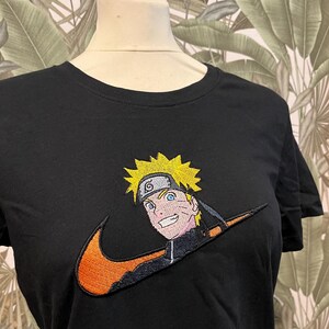 Può includere: T-shirt nera con un disegno ricamato di un personaggio dei cartoni animati con capelli gialli e occhi blu, integrato in un logo Nike arancione. Il personaggio ha una fascia nera e un'espressione sorridente.