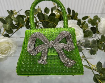 Puede incluir: Un bolso verde lima cubierto de pequeños adornos brillantes. El bolso presenta un cierre plateado en forma de lazo y un asa a juego. El bolso está sobre una superficie reflectante, con flores blancas y vegetación en el fondo.