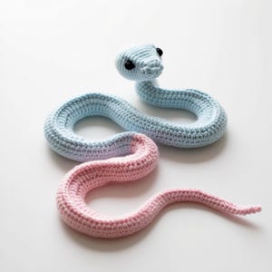 Może przedstawiać: Pluszowa zabawka w kształcie węża wykonana na szydełku, z jasnoniebieską głową i ciałem oraz różowym ogonem. Wąż ma czarne oczy i jest zwinięty w rozluźnioną pozie.