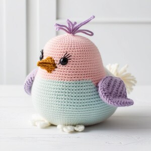 May include: A crocheted bird toy with a pink, light blue, and purple color scheme. The bird has a brown beak, black eyes, and a purple tuft on its head.