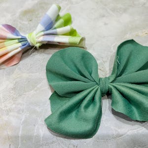 May include: Two fabric hair bows, one green and one with a plaid pattern in shades of pink, blue, and green.