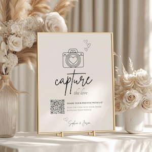 Könnte beinhalten: Ein gerahmtes Schild mit goldenem Rand und weißem Hintergrund. Das Schild zeigt eine Kamera-Grafik mit einem Herzen, die Worte "capture the love", einen QR-Code und die Namen "Sophia & Jason". Das Schild wird auf einer goldenen Staffelei angezeigt.