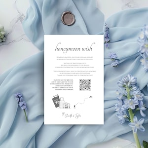Könnte beinhalten: Eine weiße Karte mit dem Text "honeymoon wish" in eleganter Schrift. Die Karte enthält einen QR-Code, Illustrationen zum Thema Reisen und eine Nachricht über die Spende für einen Flitterwochenfonds. Die Karte liegt auf einem hellblauen Stoff.
